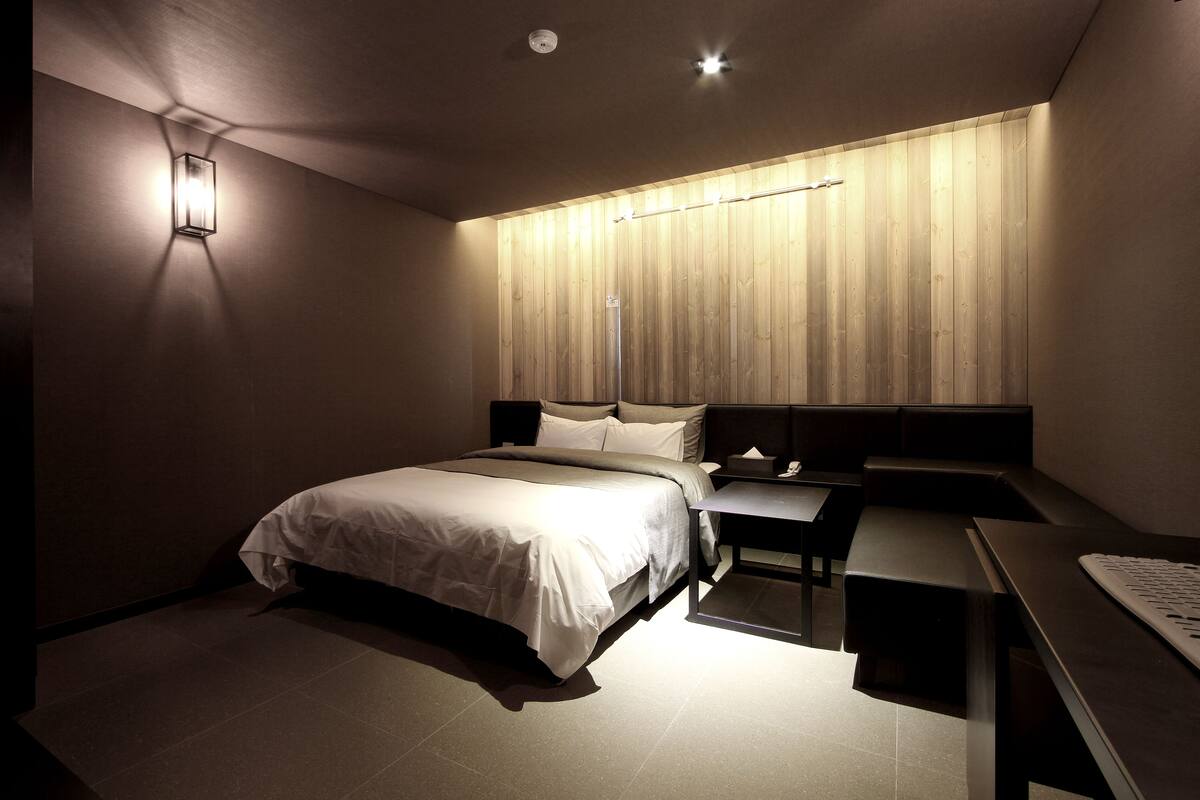 A modern hotel room with a large bed, bedside tables, a desk, and a wall-mounted light fixture.