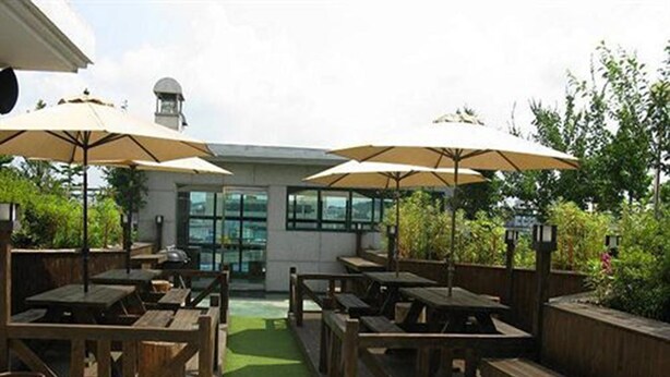 An outdoor patio with wooden tables and benches, covered by umbrellas, and a green pathway leading to a building with large windows. - Asia Tourist Hotel (Pyeongtaek)