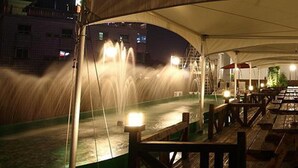 A nighttime outdoor dining area with a water fountain feature, wooden deck, and tables under a canopy. - Asia Tourist Hotel (Pyeongtaek)