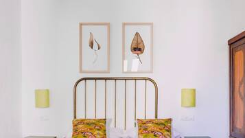 Una cama con una colcha verde, dos almohadas y un cabecero de metal. En la pared sobre la cama hay dos cuadros con motivos de hojas.