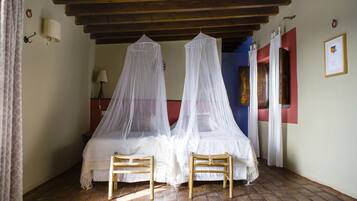 A bedroom with two beds covered by white mosquito nets, wooden chairs, a wooden ceiling, and a brick floor.