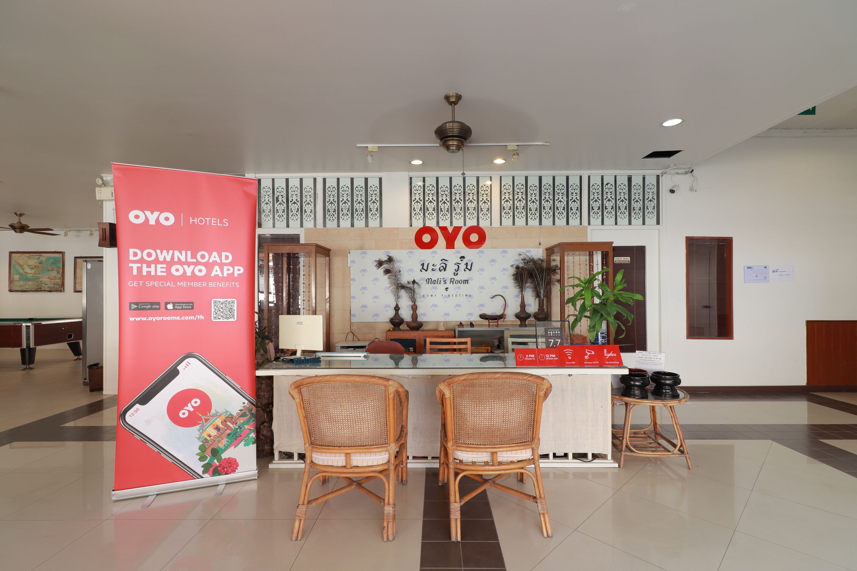 Un espace d’accueil d’hôtel comprenant un comptoir, deux fauteuils en osier, un ventilateur de plafond et un panneau publicitaire pour l’application OYO.
