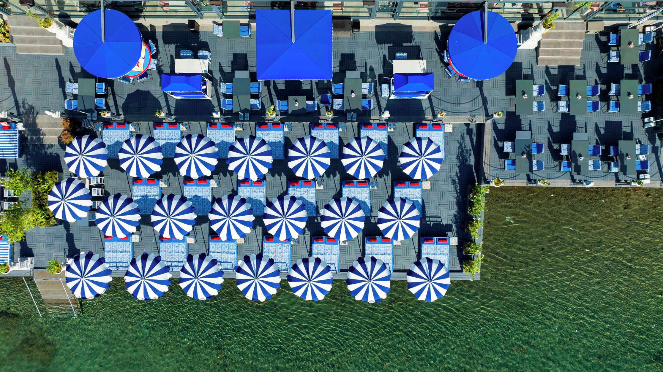 An aerial view of a hotel with multiple blue umbrellas and tables arranged in rows, a grassy area in the foreground, and a paved walkway.
