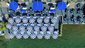 An aerial view of a hotel with multiple blue umbrellas and tables arranged in rows, a grassy area in the foreground, and a paved walkway. - Grand Yazici Club Turban (Marmaris)