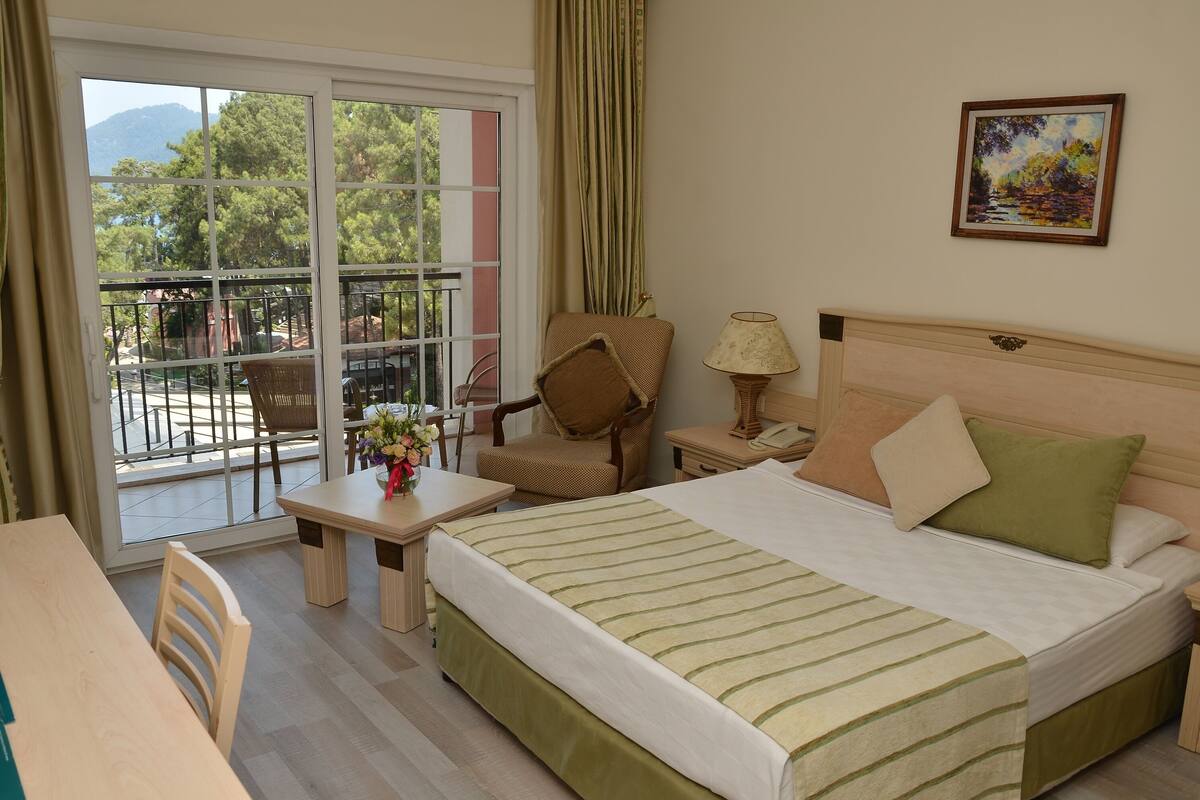A hotel room with a bed, a chair, a table, and a balcony with a view of trees and mountains.