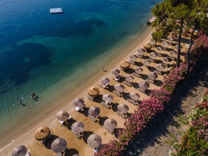 An aerial view of a beach with rows of sun loungers and thatched umbrellas, clear blue water, and a boat in the distance. - Grand Yazici Club Turban (Marmaris)