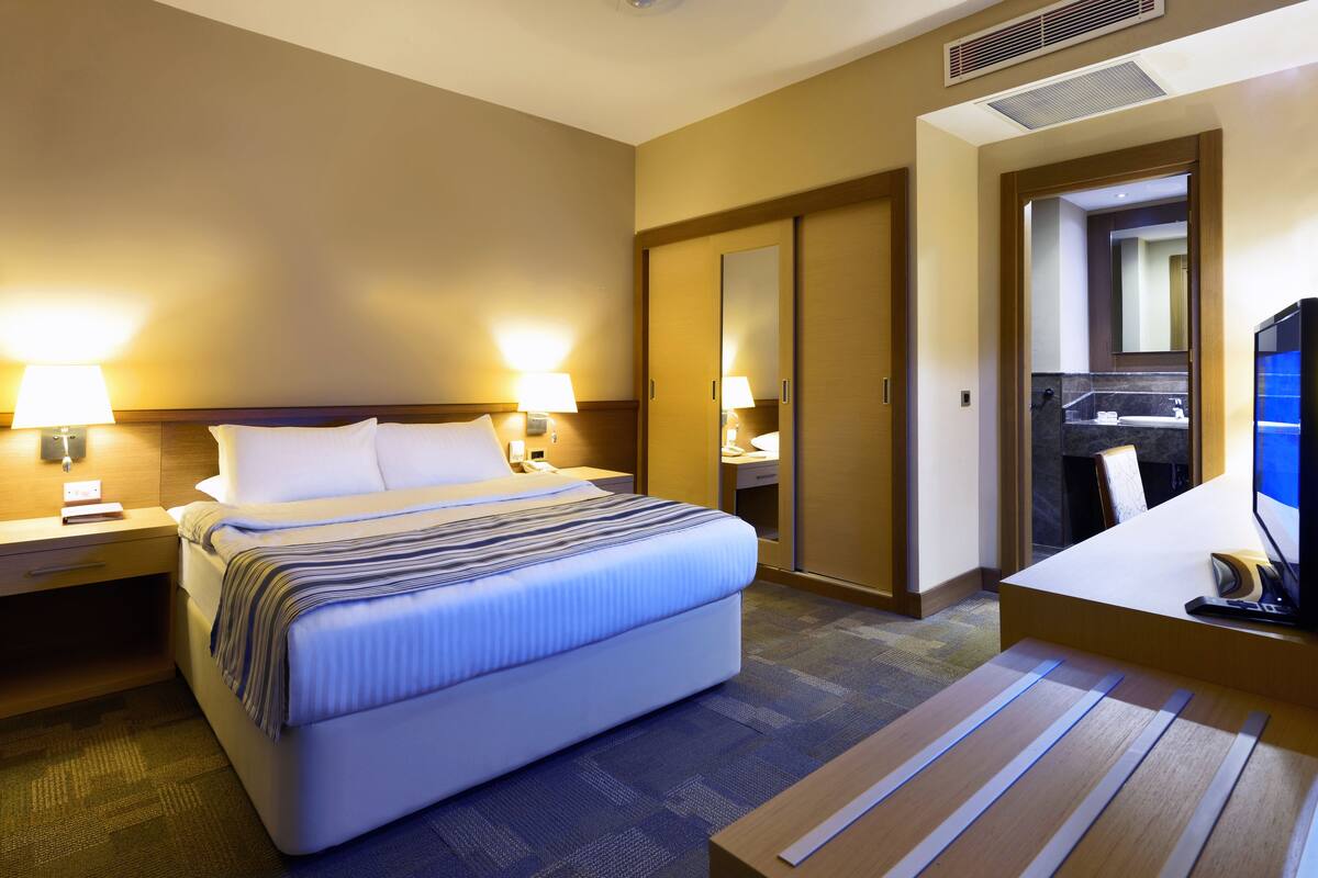 A hotel room with a bed, bedside lamps, a desk with a computer, and a bathroom visible through an open door.