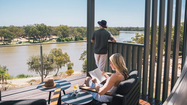 A balcony with a view of a river, a person reading a book, and another person standing by the railing. - The Frames (Paringa)
