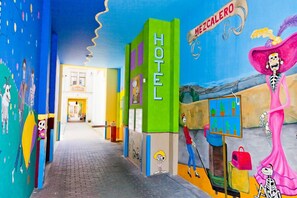 Un couloir d’hôtel aux couleurs vives, avec un bâtiment vert sur lequel on peut lire l’inscription « HÔTEL » et une fresque représentant des squelettes et une femme en robe rose.