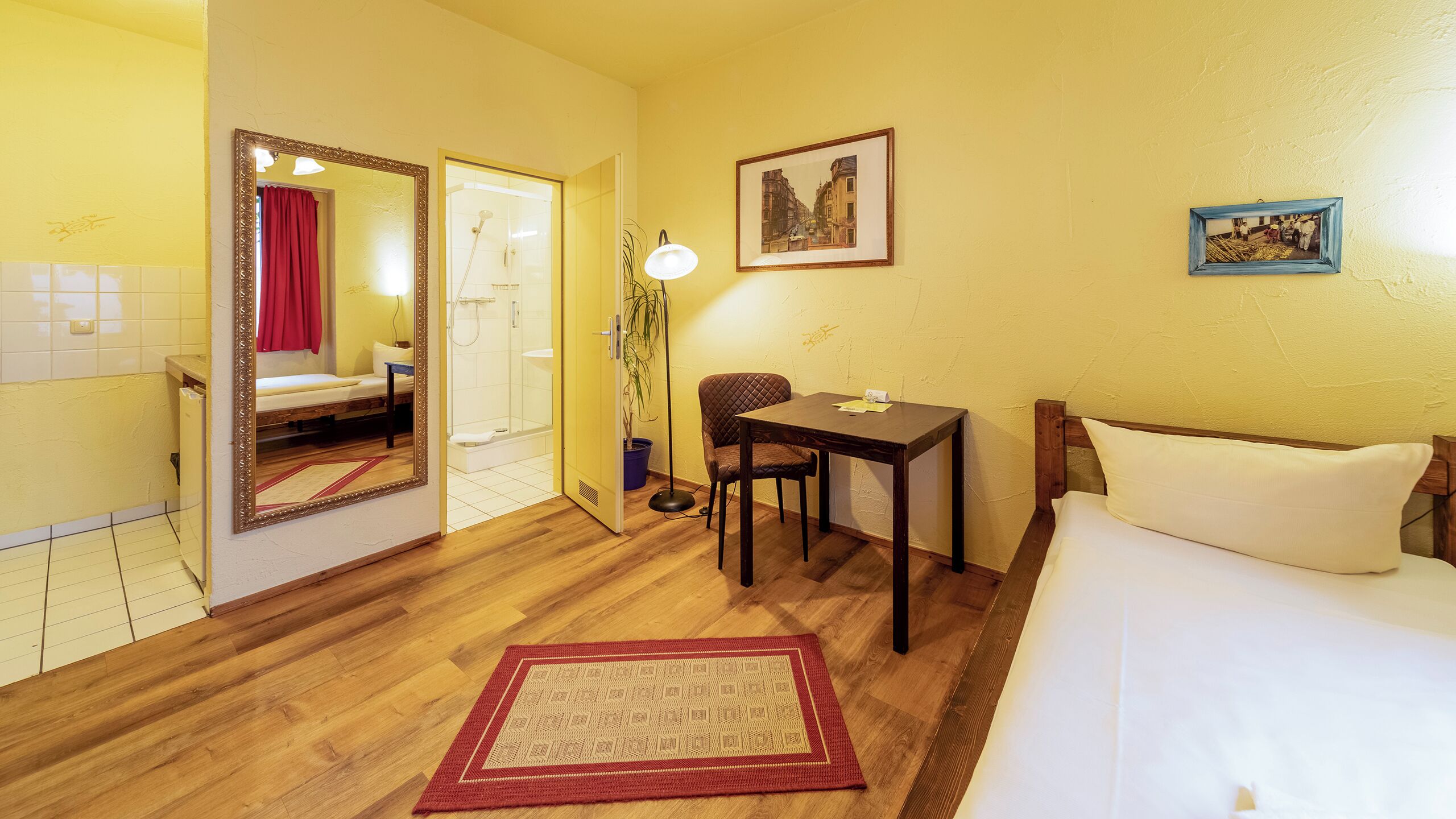 A hotel room with a bed, a desk, a chair, a mirror, a shower, and a small table with a lamp.