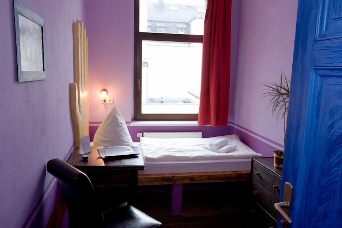 Een kleine kamer met een enkel bed, een bureau en een stoel. Er is een raam met uitzicht op gebouwen.