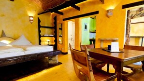 Een kamer met een bed, een houten eettafel en stoelen. Achtergrond toont een keukenhoek.