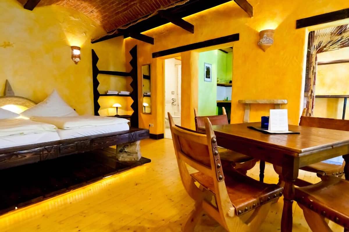 Een kamer met een bed, een houten eettafel en stoelen. Achtergrond toont een keukenhoek.