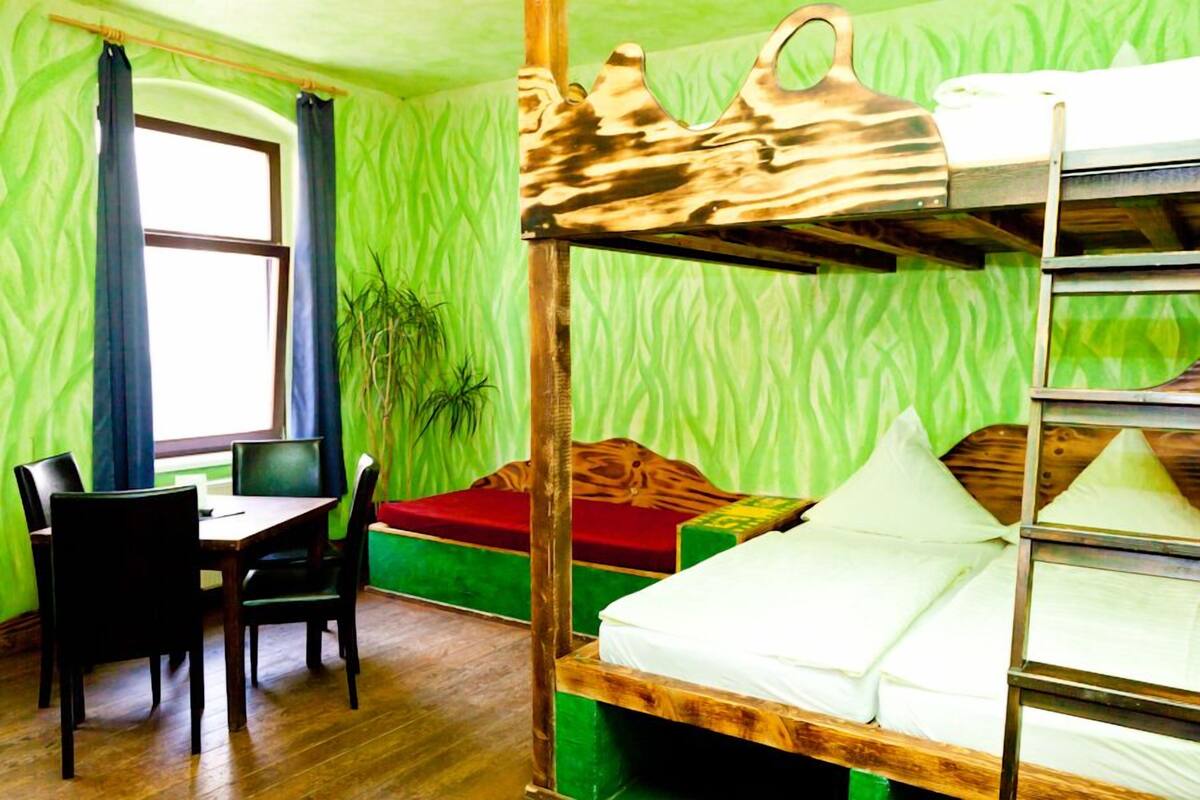 Een kamer met stapelbedden, een bureau met stoel, een kleine tafel en een raam met gordijnen.