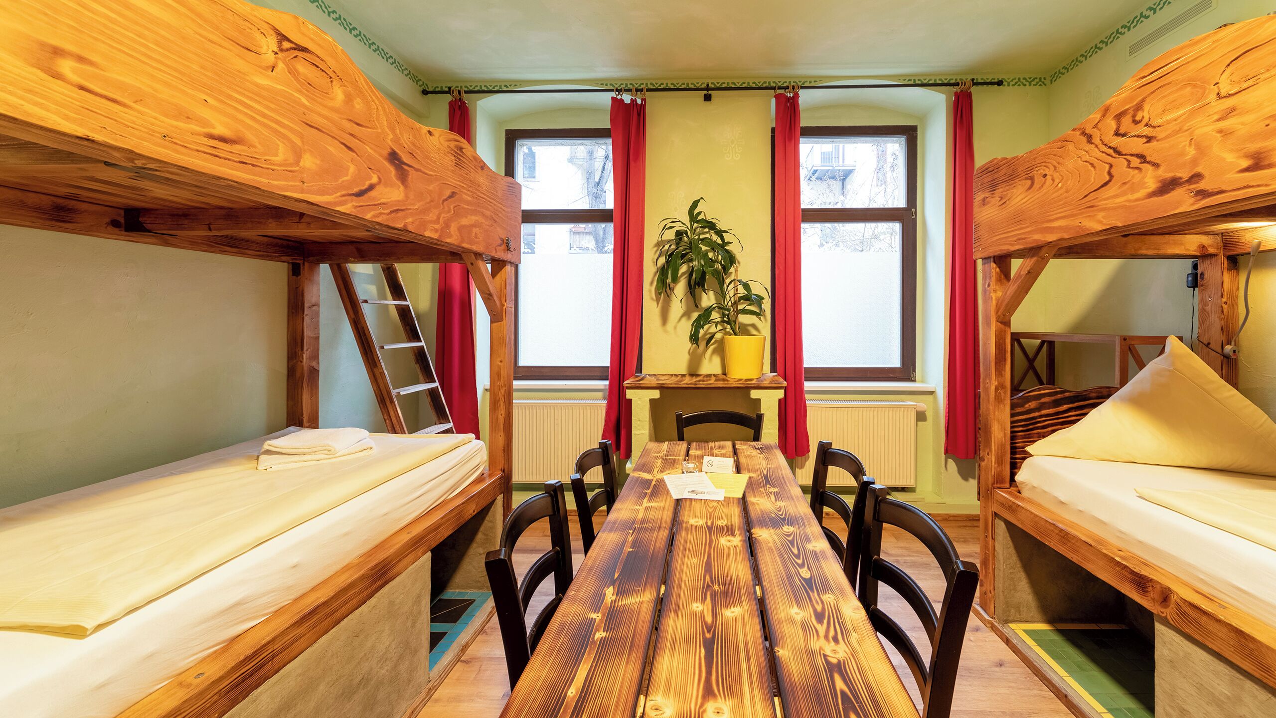 A room with bunk beds, a wooden table, chairs, a plant, and a window with red curtains.
