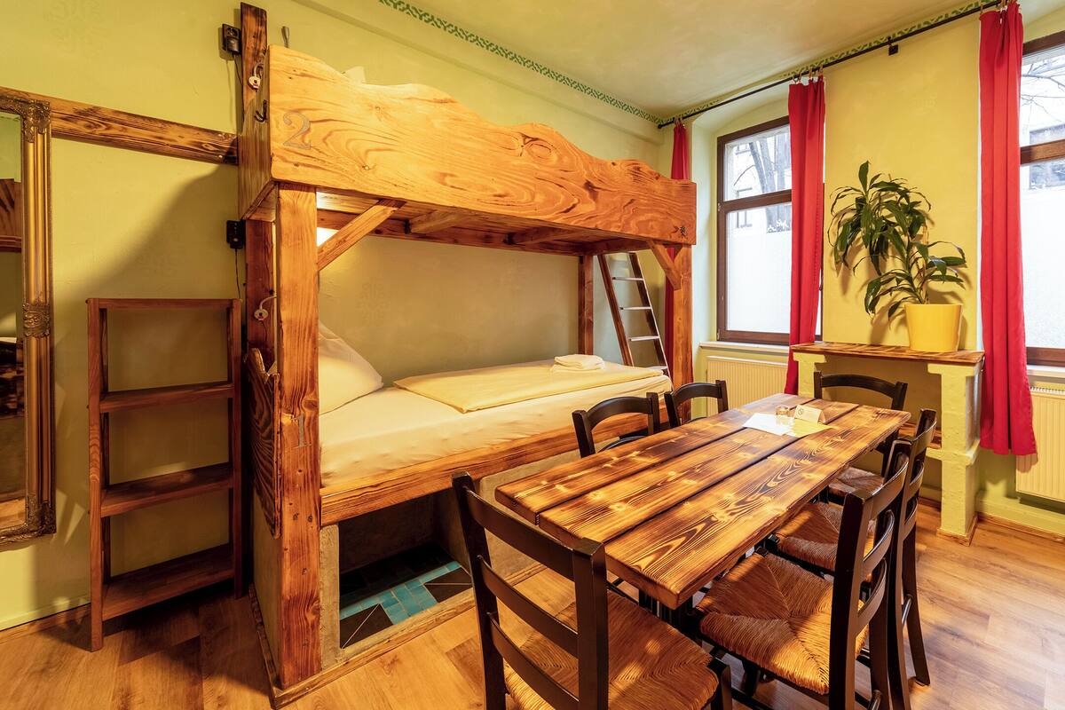 Een stapelbed met een houten ladder, een houten tafel met stoelen en een potplant bij een raam met rode gordijnen.