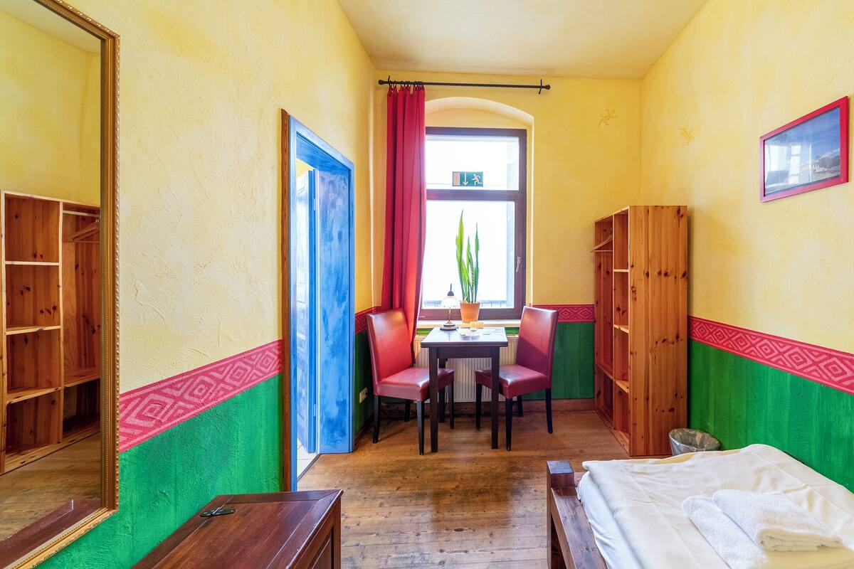Een smalle hotelkamer met een bed, twee stoelen, een tafel en een raam met gordijnen.
