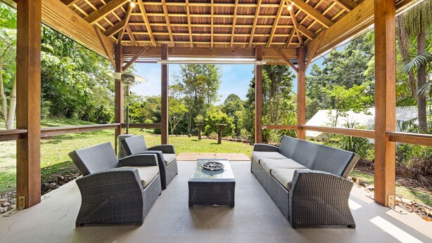 Un patio exterior con mobiliario de mimbre y una mesa de centro, rodeado por una pérgola de madera con vista a un jardín exuberante.