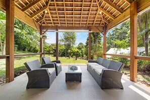 Un patio exterior con mobiliario de mimbre y una mesa de centro, rodeado por una pérgola de madera con vista a un jardín exuberante.