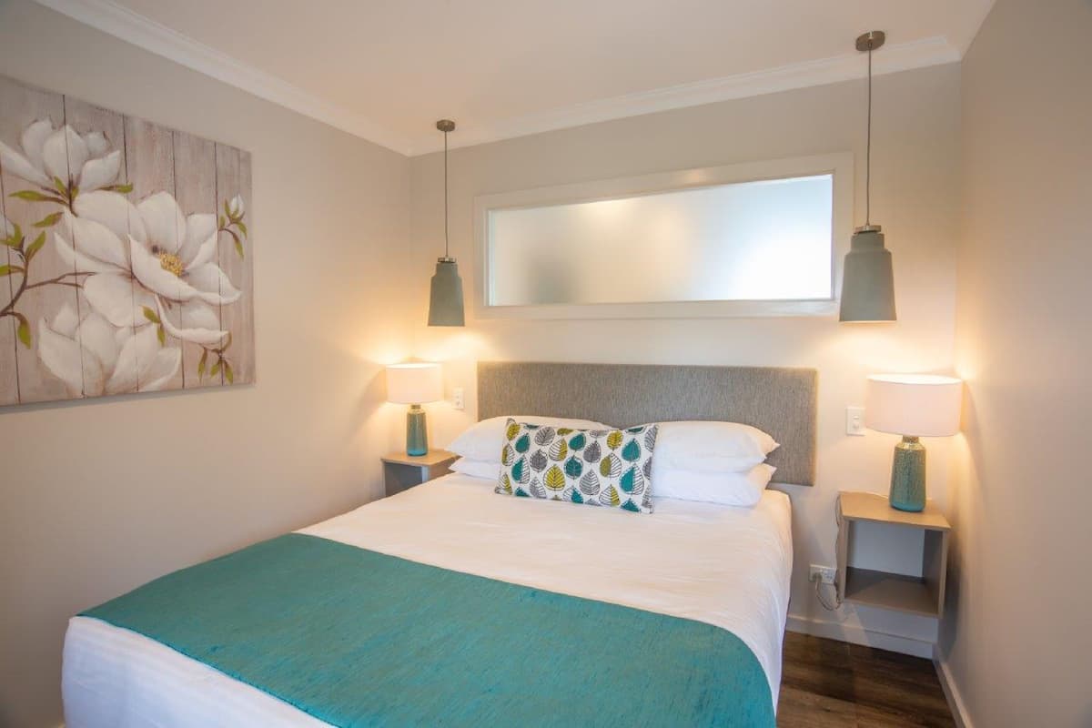 Una cama bien hecha con un edredón verde azulado, dos lámparas de noche, un cuadro floral grande y un espejo grande sobre la cama.