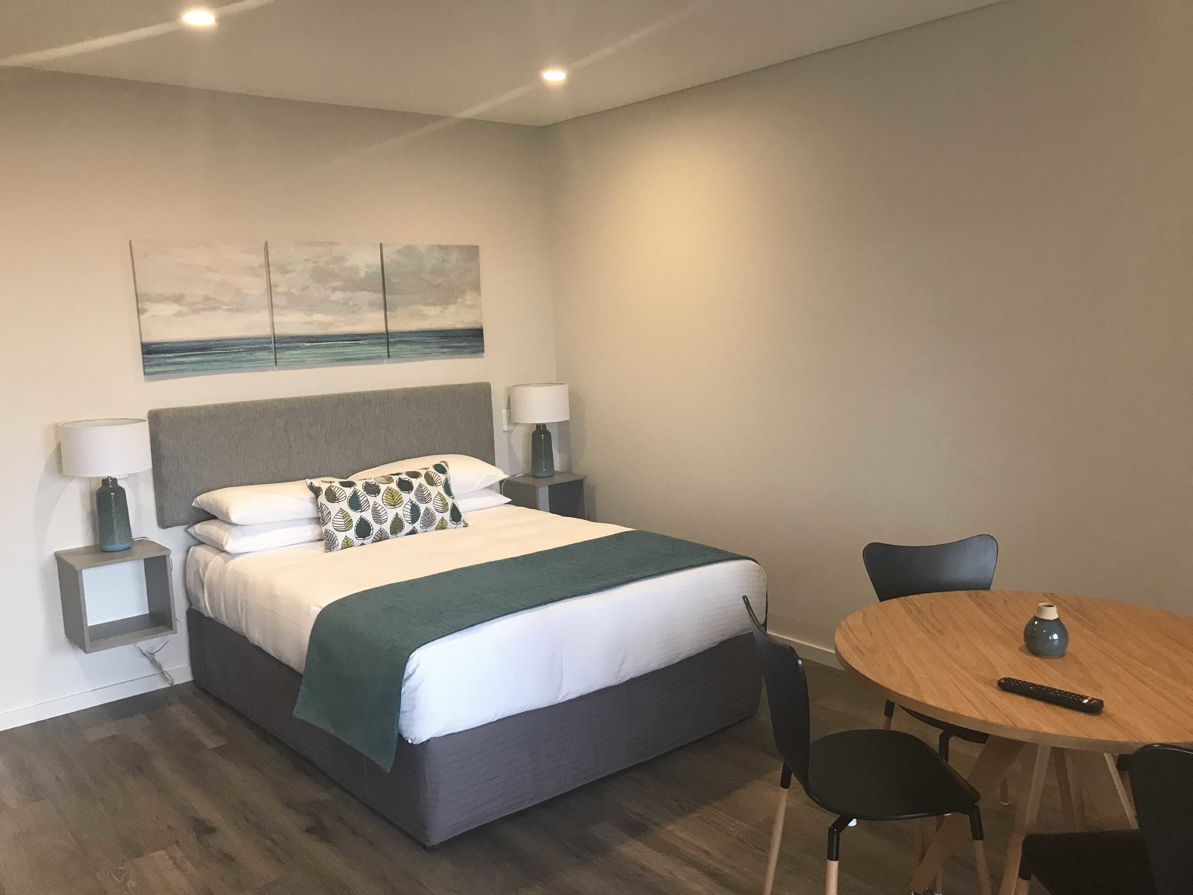 A modern hotel room with a bed, bedside lamps, a table with chairs, and a painting on the wall.