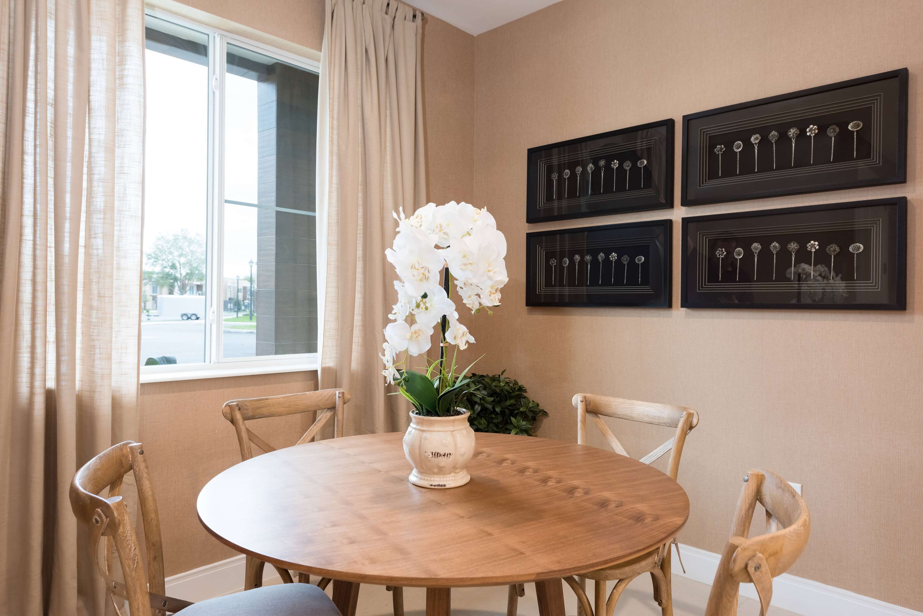 A dining area with a round wooden table, four wooden chairs, a vase with white flowers, and four framed floral artworks on the wall.