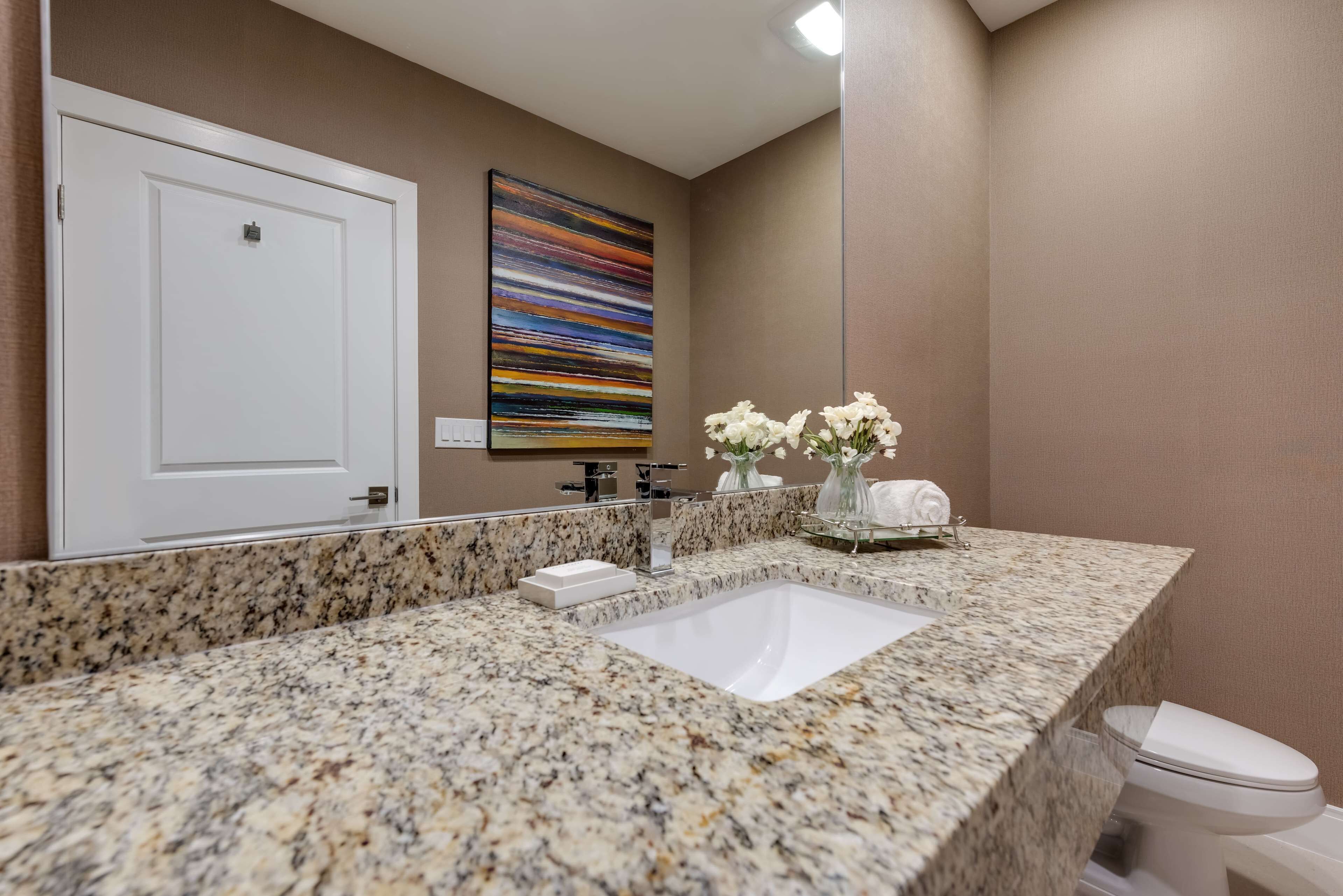 Une salle de bain avec un comptoir en granit, un lavabo blanc, un miroir, une porte et un vase contenant des fleurs blanches.