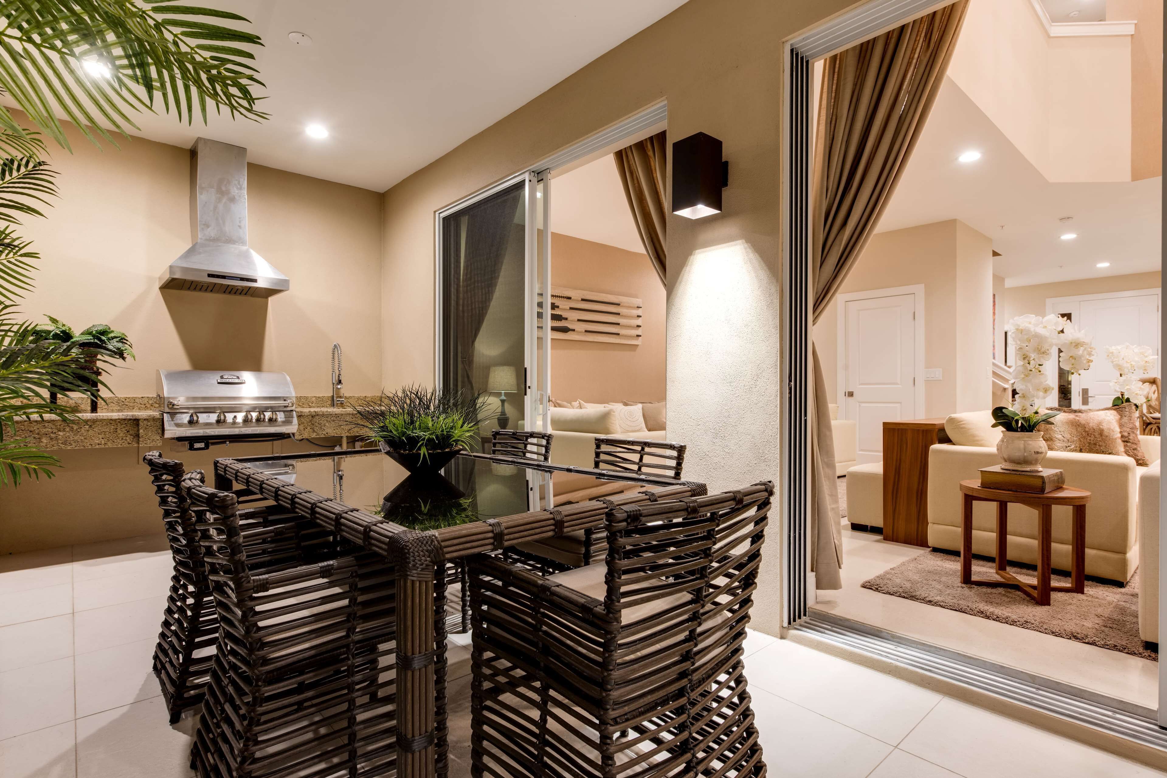 A modern kitchen with a built-in grill, dining area with wicker chairs, and a living room with a sofa and decorative plants.