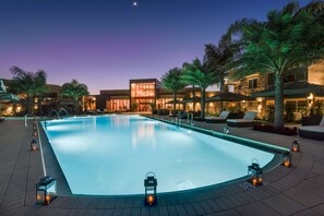 Une piscine dotée d’une terrasse en bois, entourée de palmiers et de chaises longues, avec des bâtiments en arrière-plan.