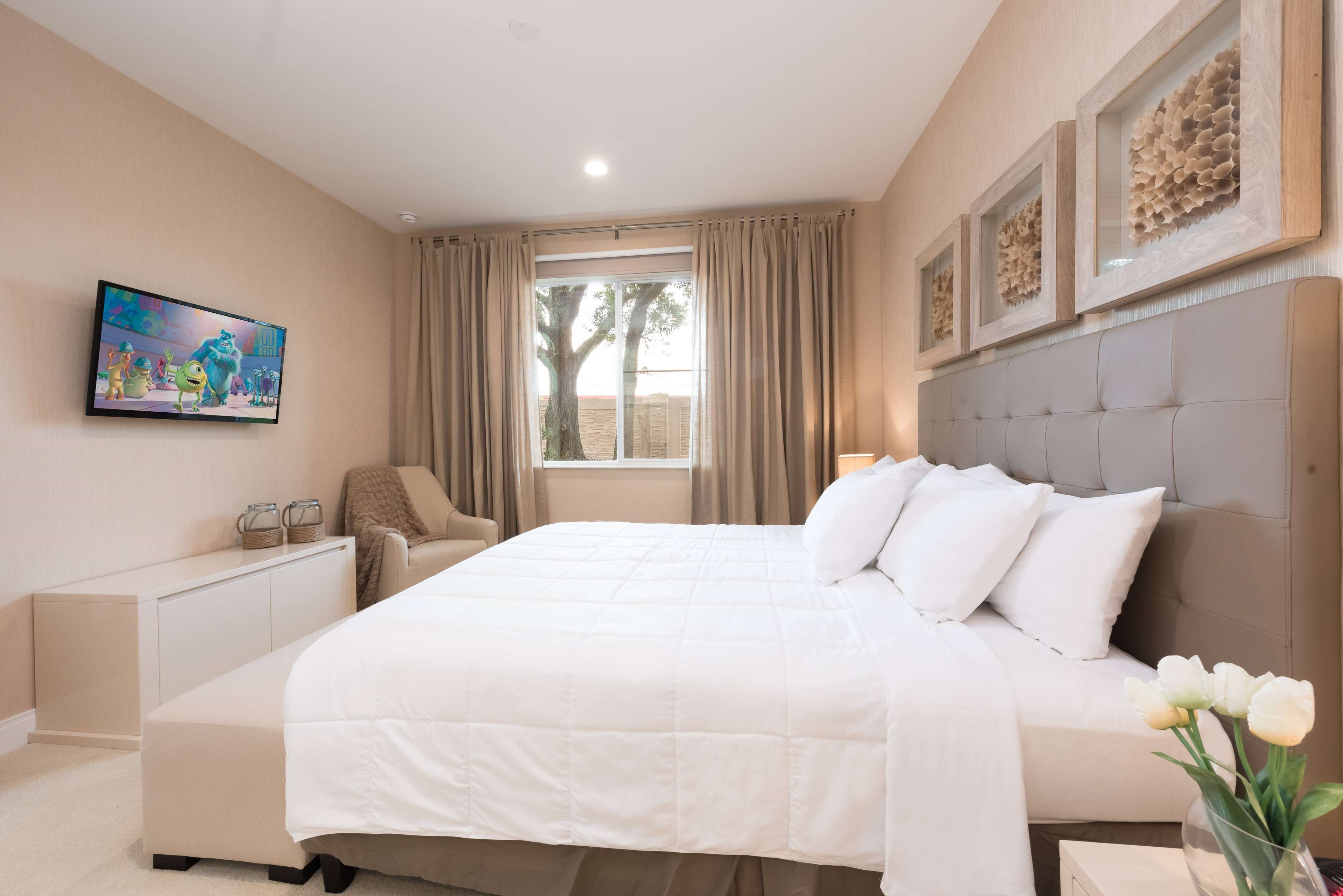 A bedroom with a large bed, a bedside table, a chair, a desk, a window with curtains, and a wall-mounted TV.