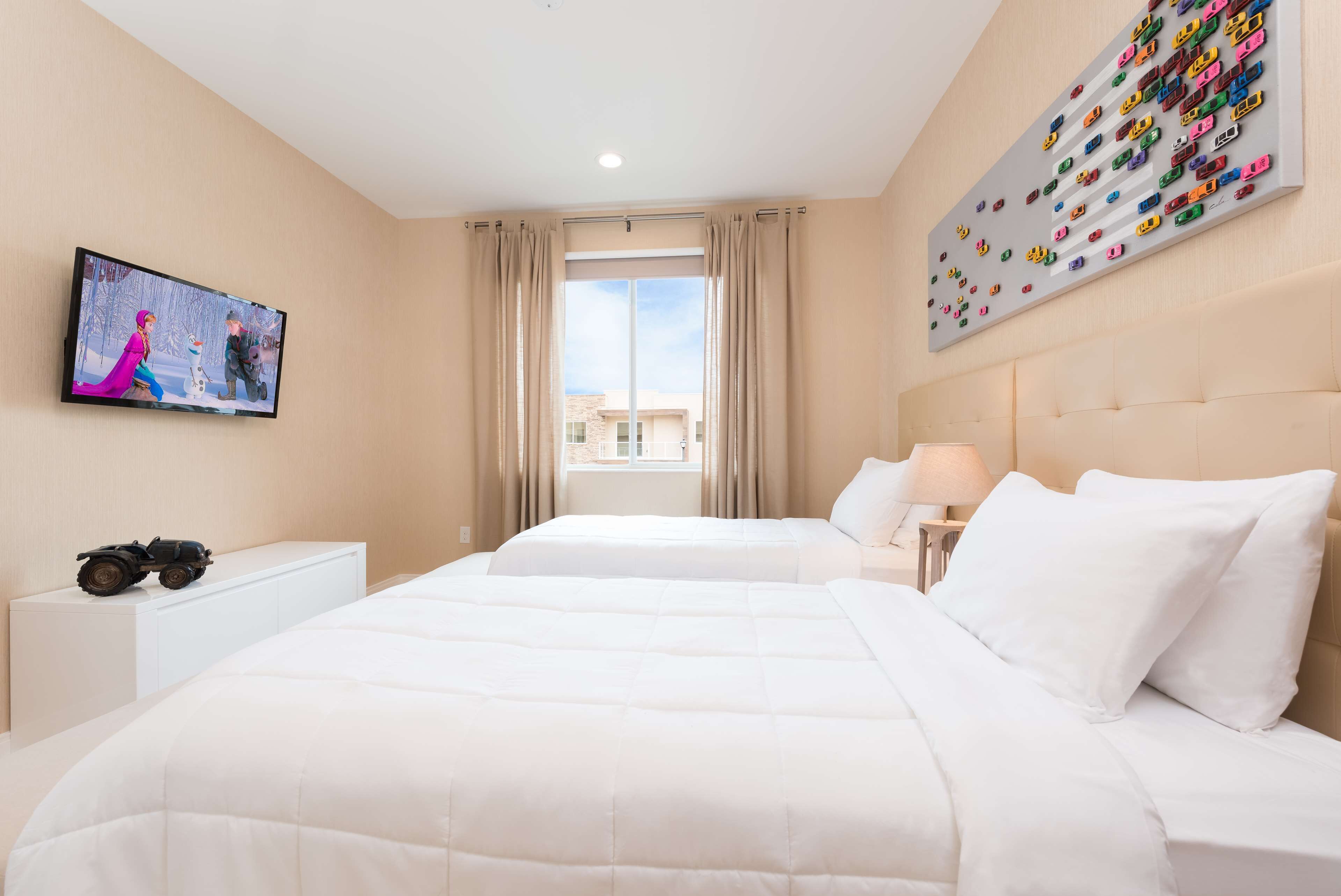 Habitación de hotel con dos camas, un televisor, un cuadro con coches de juguete y una ventana con cortinas.