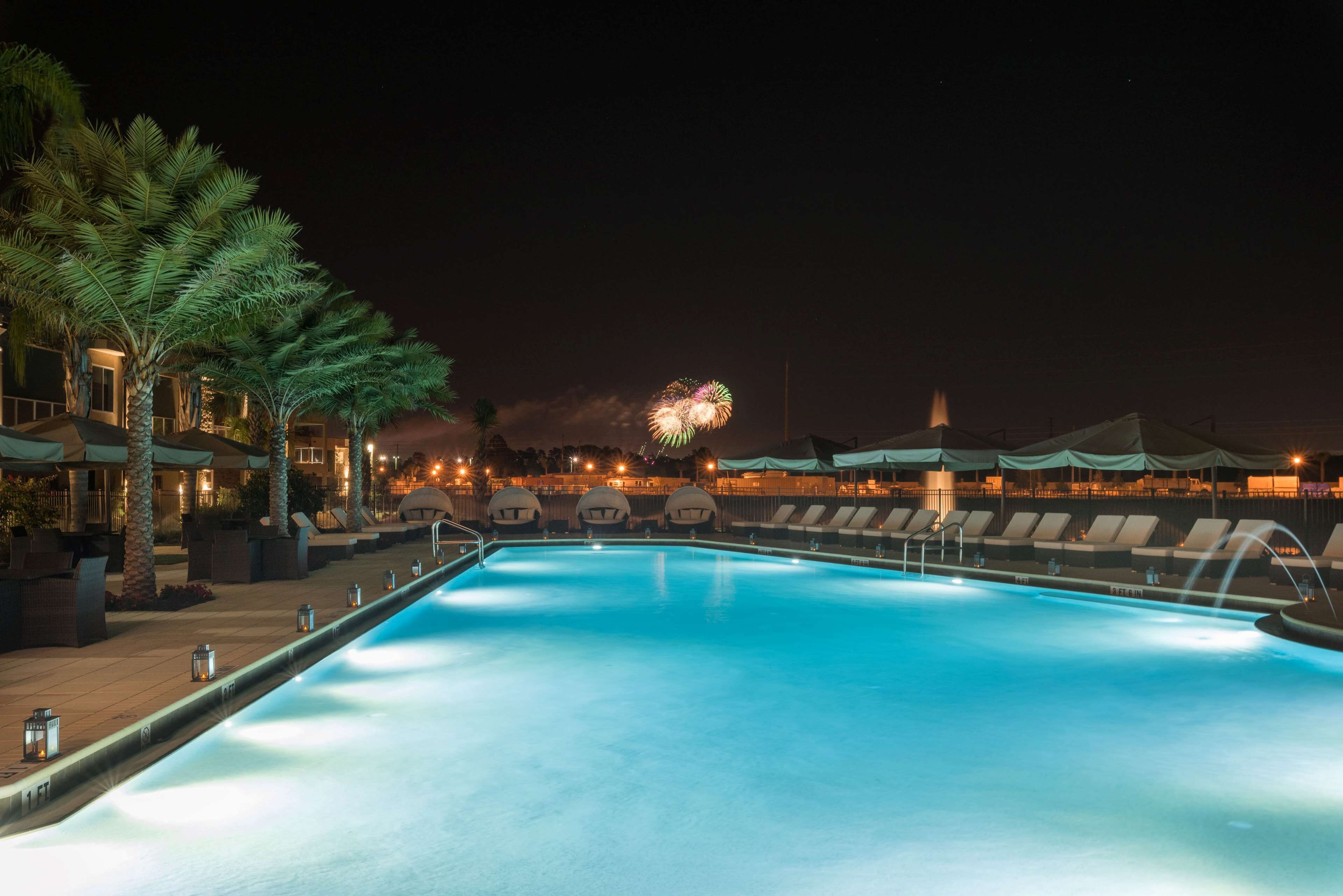 Una piscina con sillas de descanso y palmeras, con fuegos artificiales al fondo.