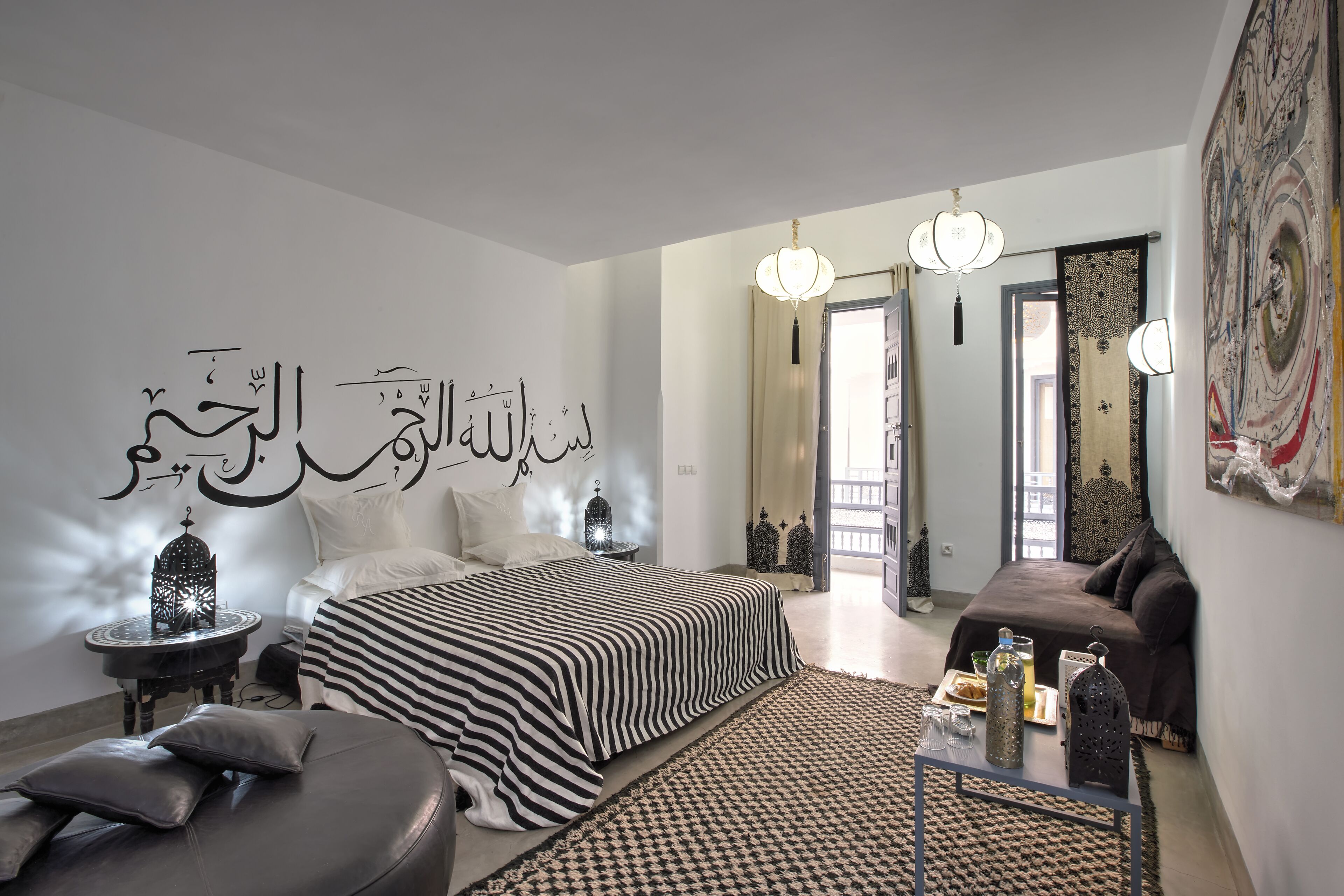 A modern bedroom with a large bed, a sofa, a small table with decorative items, and a wall with Arabic calligraphy.