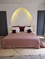 Una cama con colcha a rayas, una pared decorativa con diseño geométrico y una pequeña estantería con un objeto decorativo.