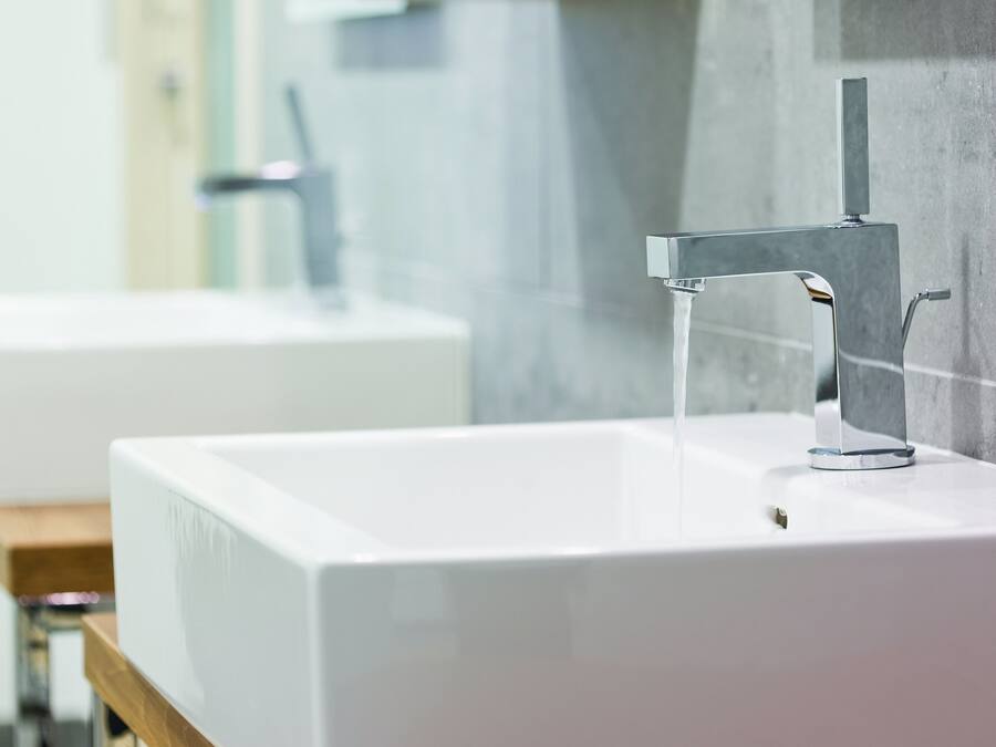 A modern bathroom with a white sink, chrome faucet, and a wooden shelf above it.