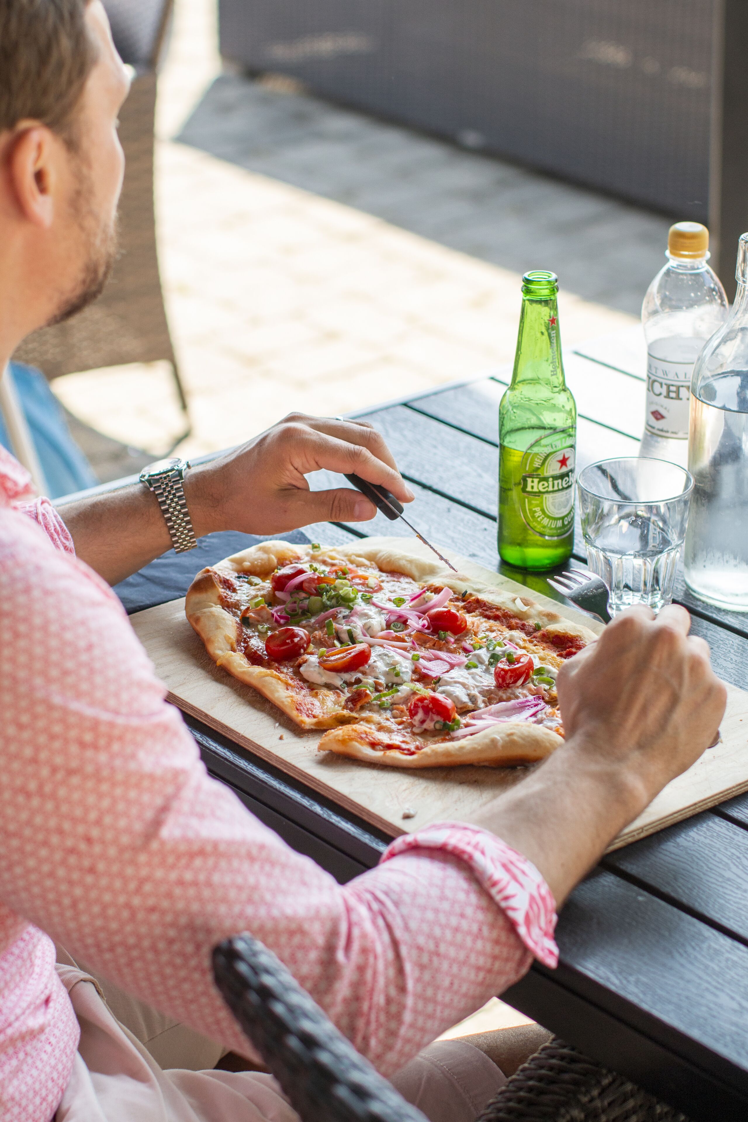 Eine Person sitzt an einem Holztisch mit einer Pizza, einer grünen Flasche und einem Glas Wasser.