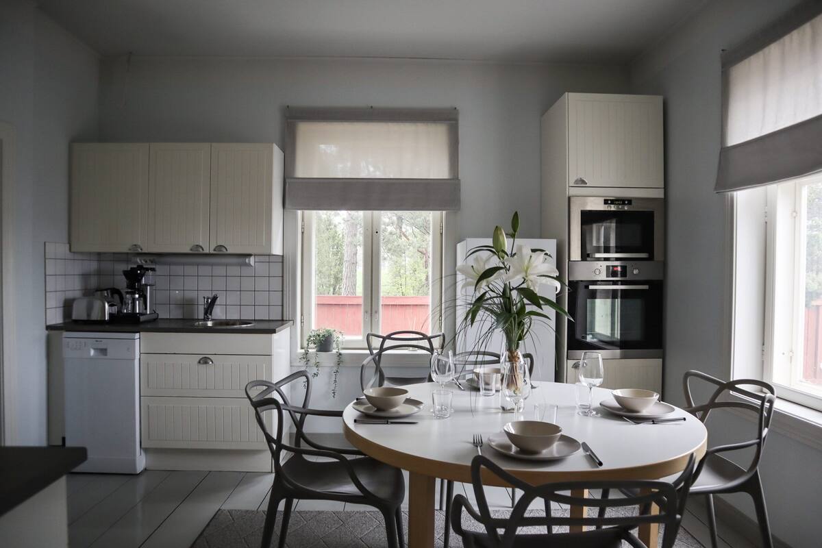 Une cuisine moderne avec des meubles blancs, une table à manger pour quatre personnes et une grande fenêtre qui laisse entrer la lumière naturelle.