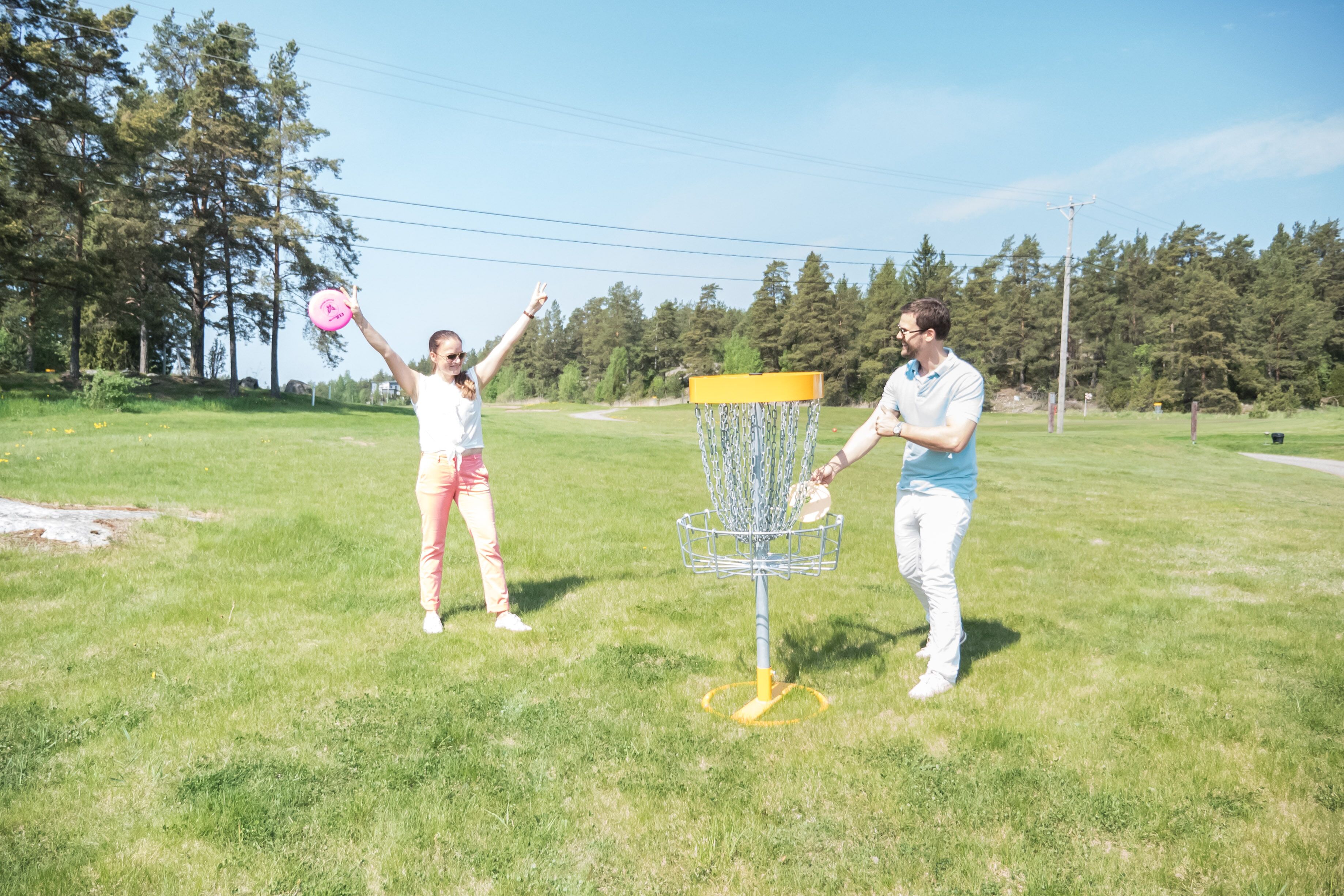 Two people playing frisbee in a grassy field with a disc golf basket.