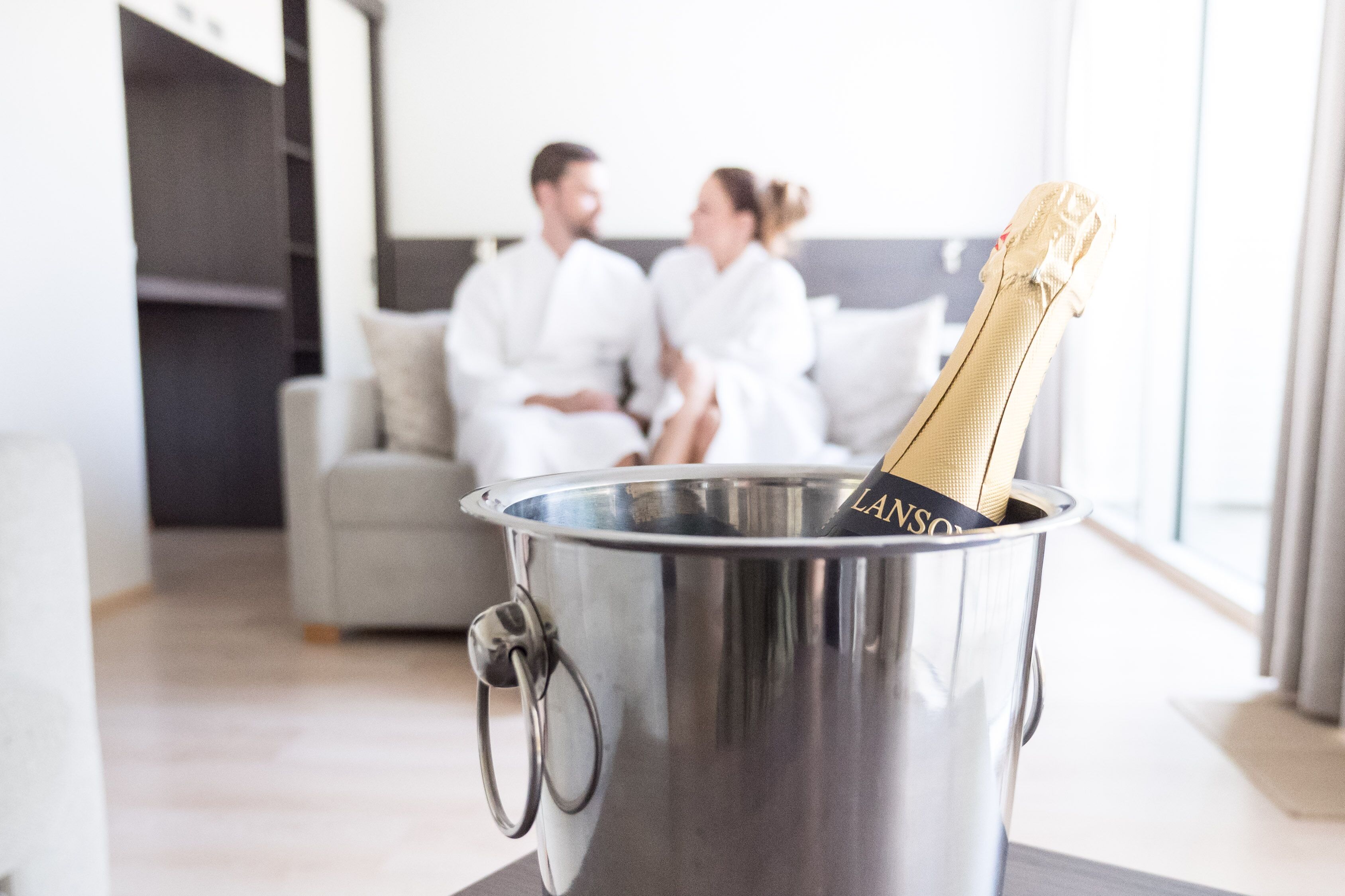 Un balde metálico con una botella de champán, un sofá y una persona con bata de baño al fondo.