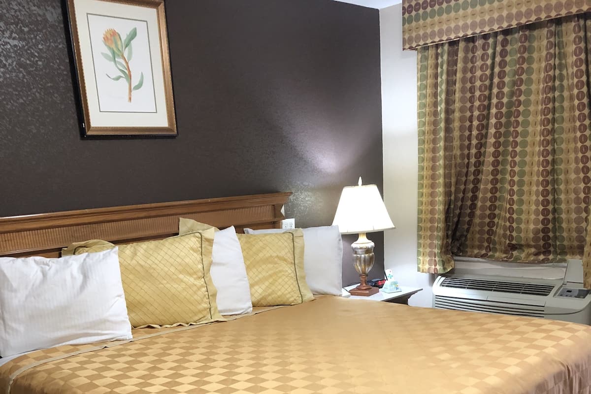 A hotel room with a large bed, a nightstand with a lamp, a wall-mounted air conditioner, and a window with curtains.