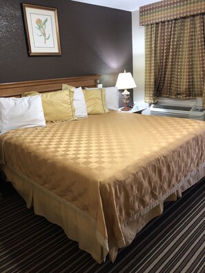 A hotel room with a large bed, a nightstand with a lamp, a wall-mounted air conditioner, and a window with curtains.