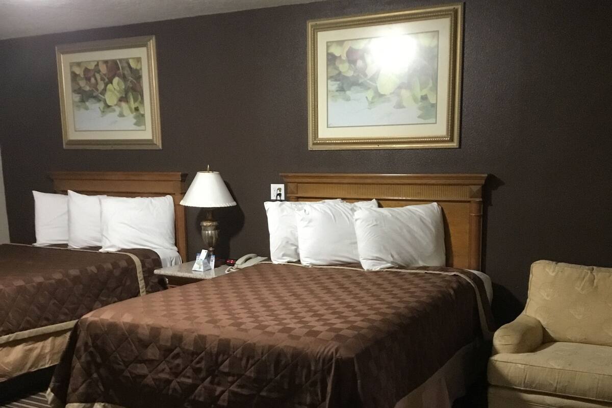 A hotel room with two beds, a chair, a nightstand with a lamp, and two framed pictures on the wall.