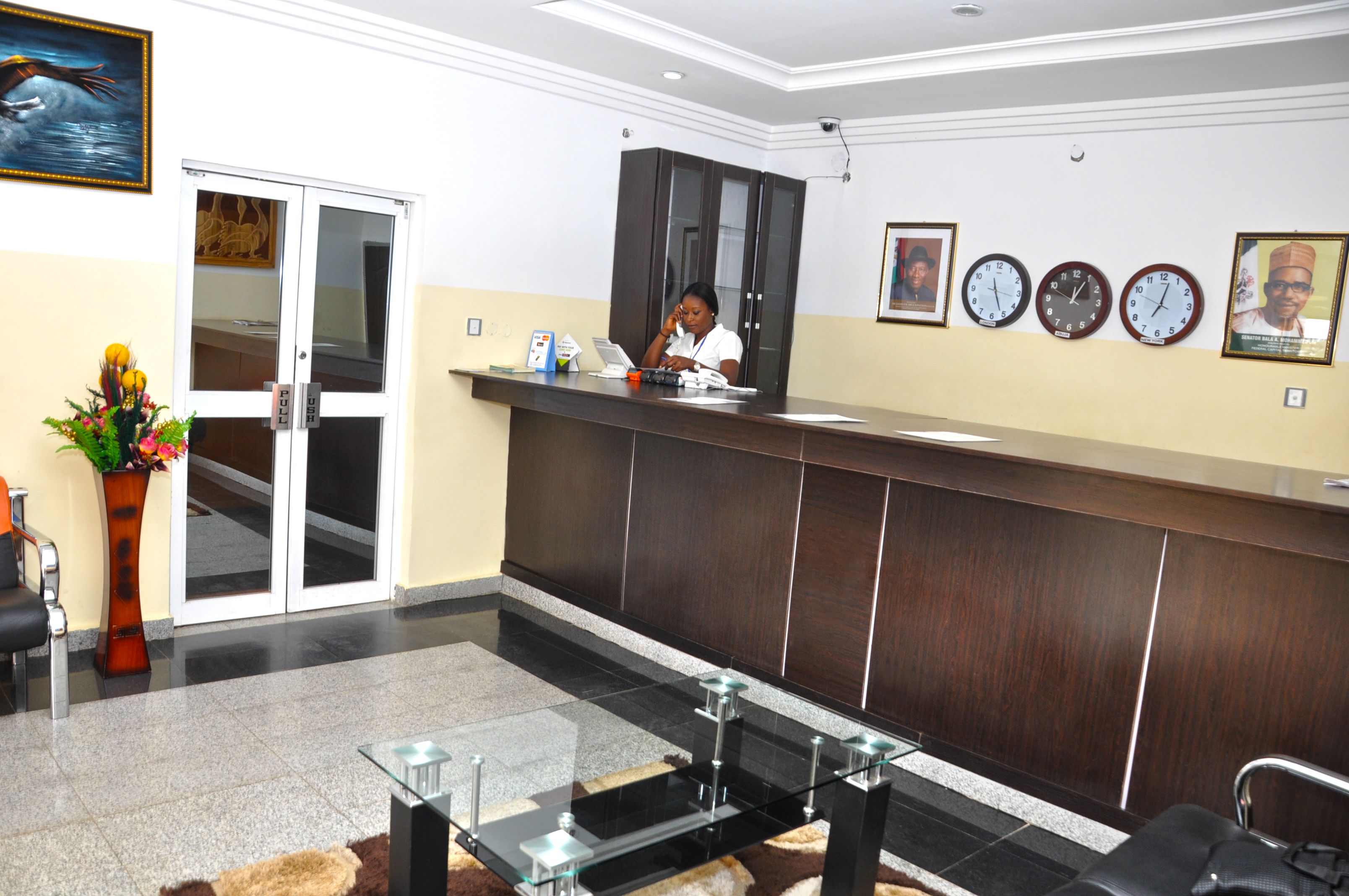 A reception area with a receptionist, a reception desk, a glass table, chairs, a vase with flowers, and framed pictures on the wall.