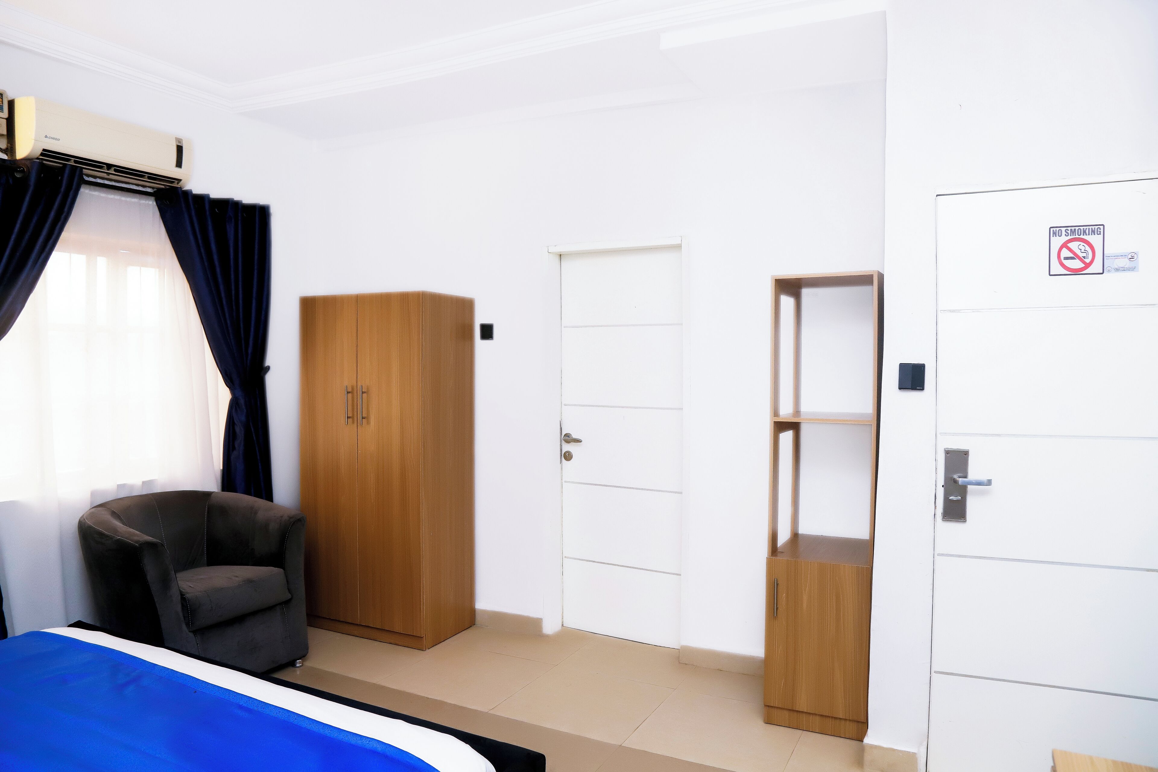 A room with a bed, a chair, a wardrobe, a door, and a shelf unit.
