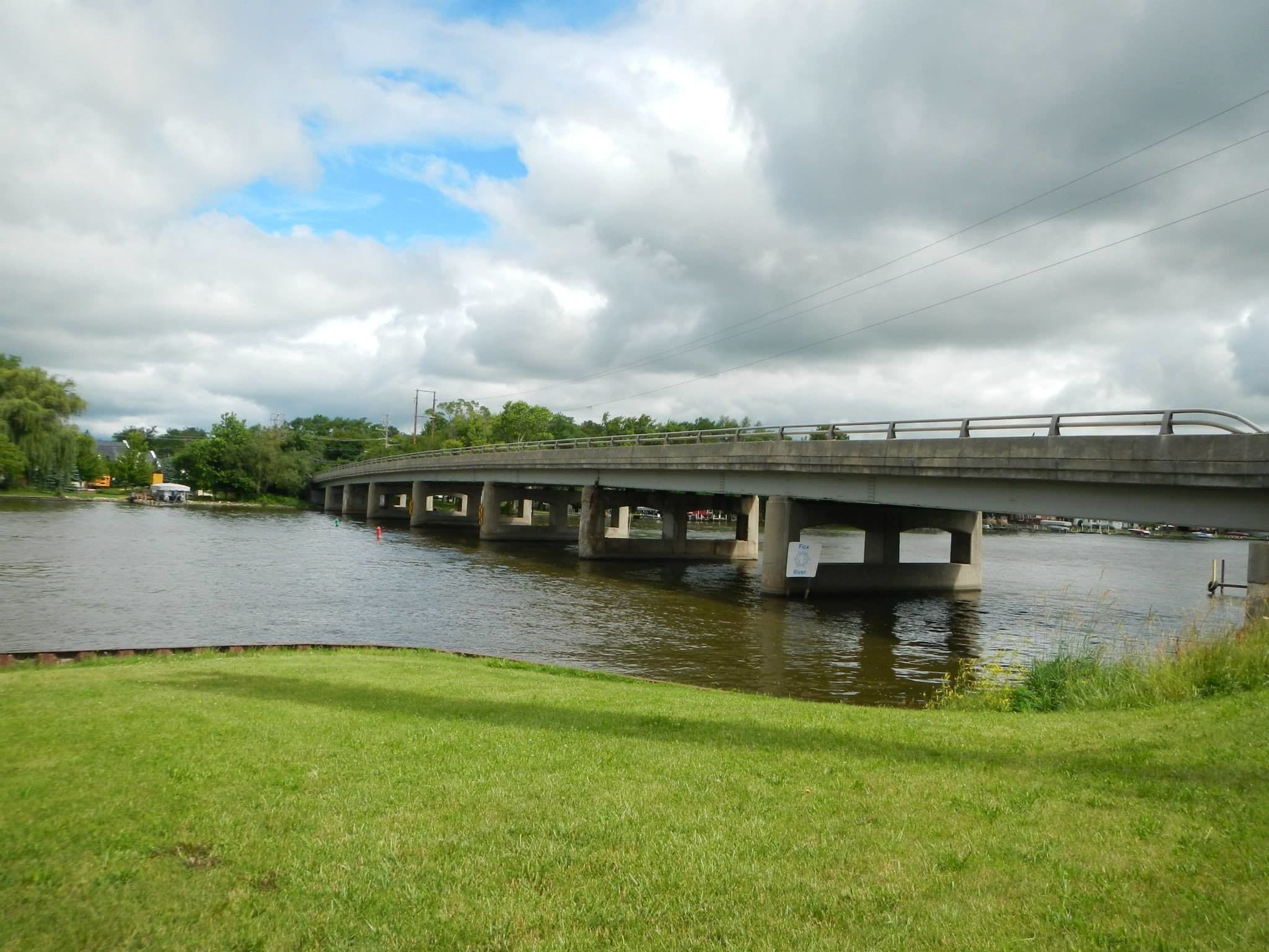 A concrete bridge over a river with grassy banks and trees in the background.