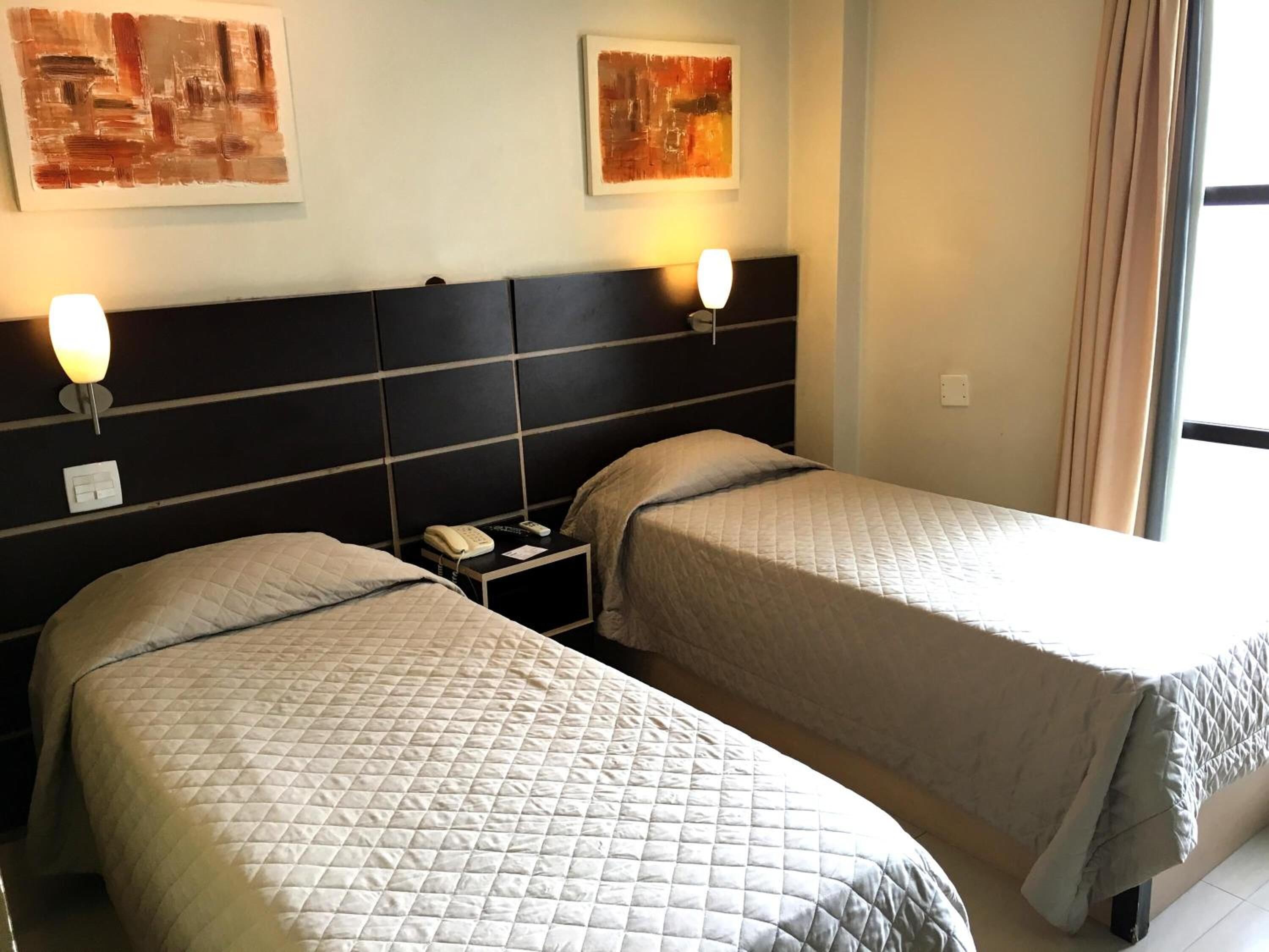Et hotelværelse med to senge, hver med et bølget sengetæppe, et natbord med lampe og en telefon ophængt på væggen.
