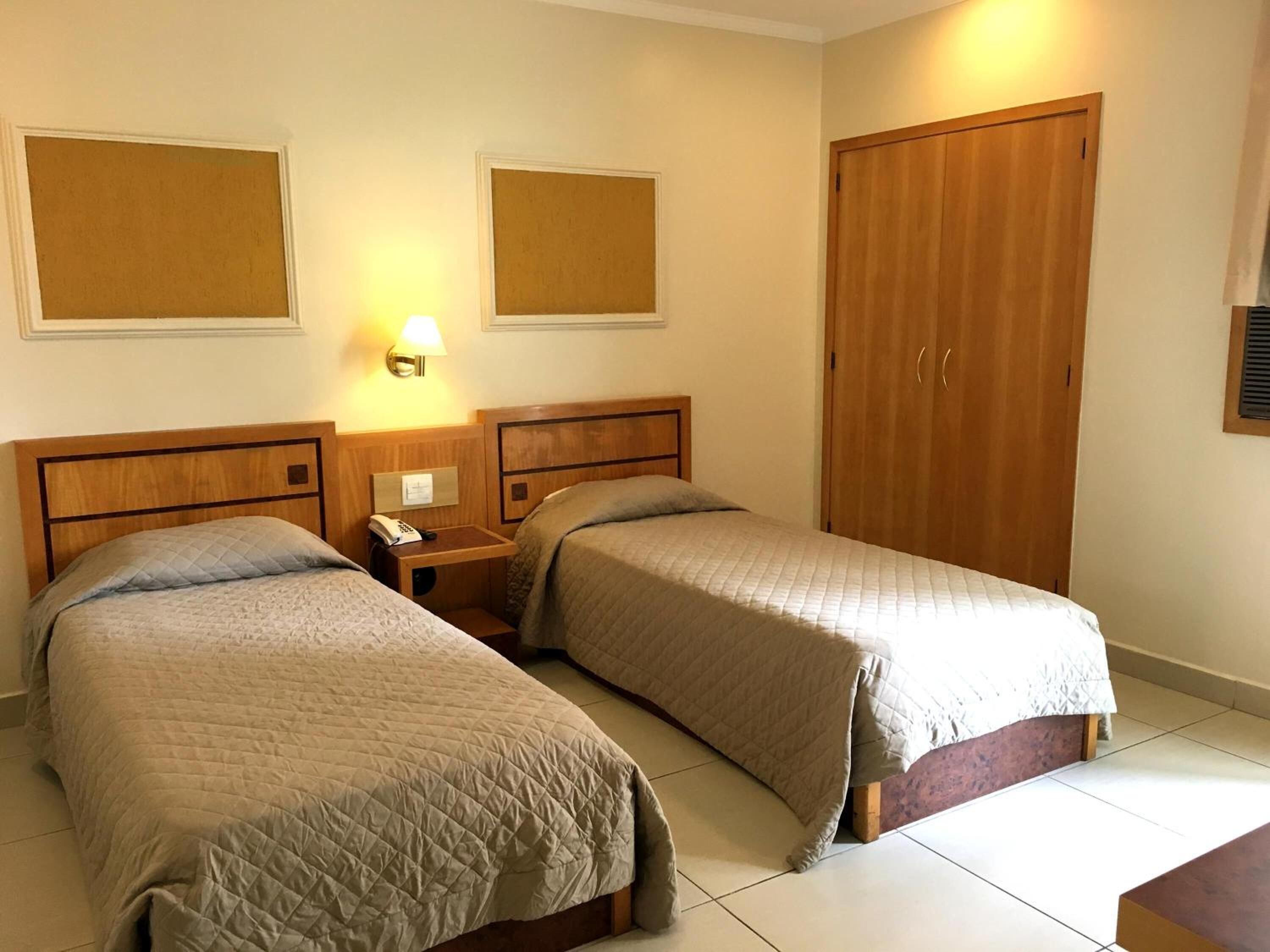 A hotel room with two single beds, a wooden wardrobe, a bedside table with a lamp, and a wall-mounted telephone.