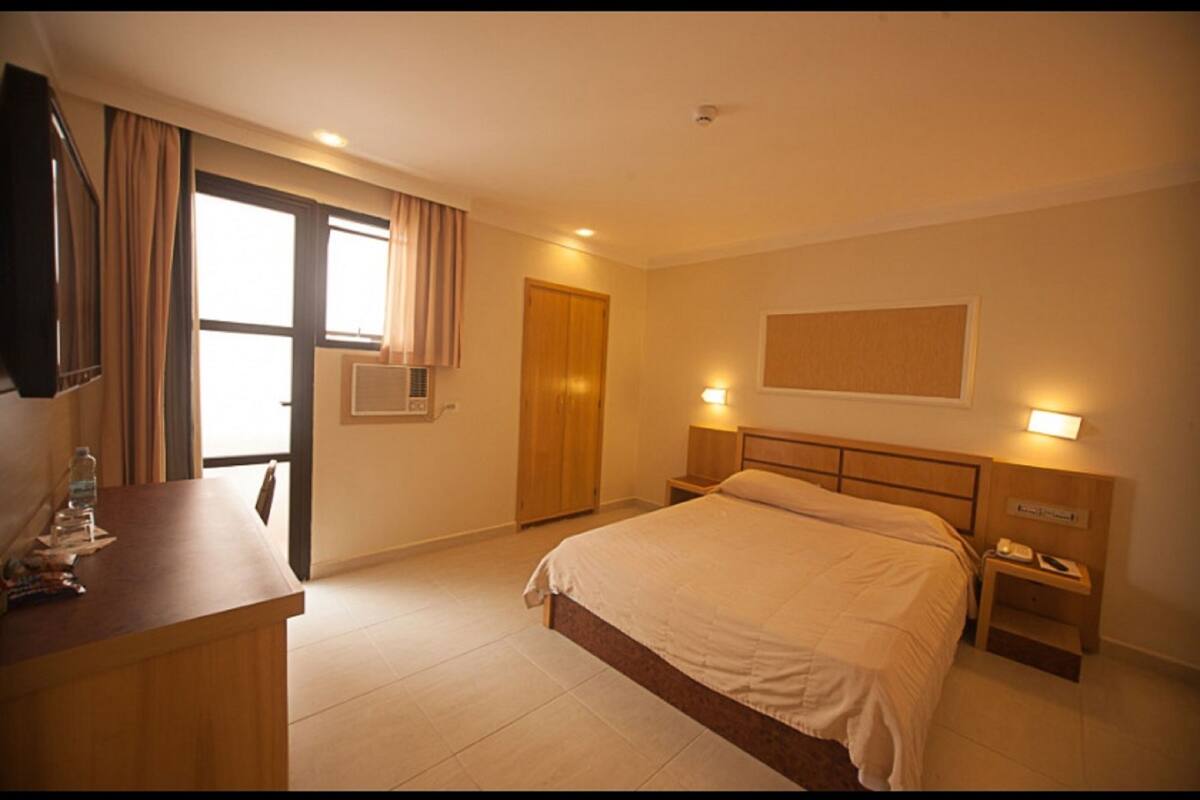 A hotel room with a bed, bedside tables, a desk, a television, and a window with curtains.