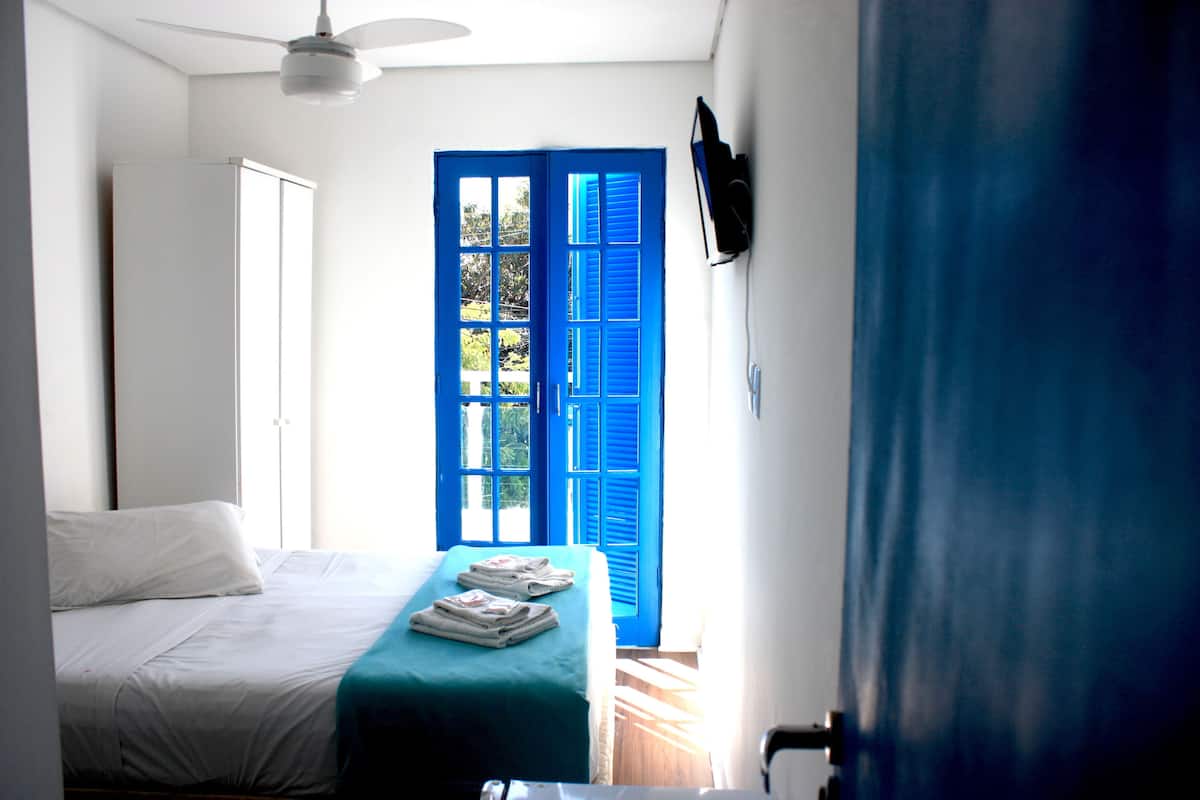 A bedroom with a bed, a ceiling fan, a window with blue shutters, and a wall-mounted TV.