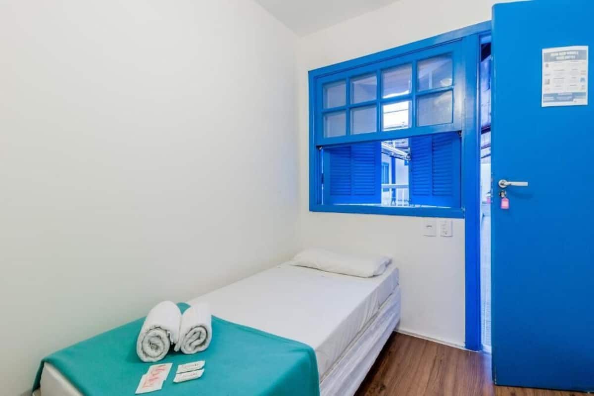 A single bed with a teal bedspread, white pillows, and rolled towels. A blue door with a window is visible, and a small sign is attached to the door.